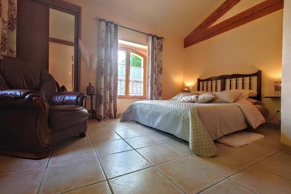 Chambre rustique et confortable avec carrelage en terre cuite, plafond aux poutres apparentes et lumi&egrave;re naturelle chaude filtrant &agrave; travers les rideaux des fen&ecirc;tres, parfaite pour un week-end de chasse en France.