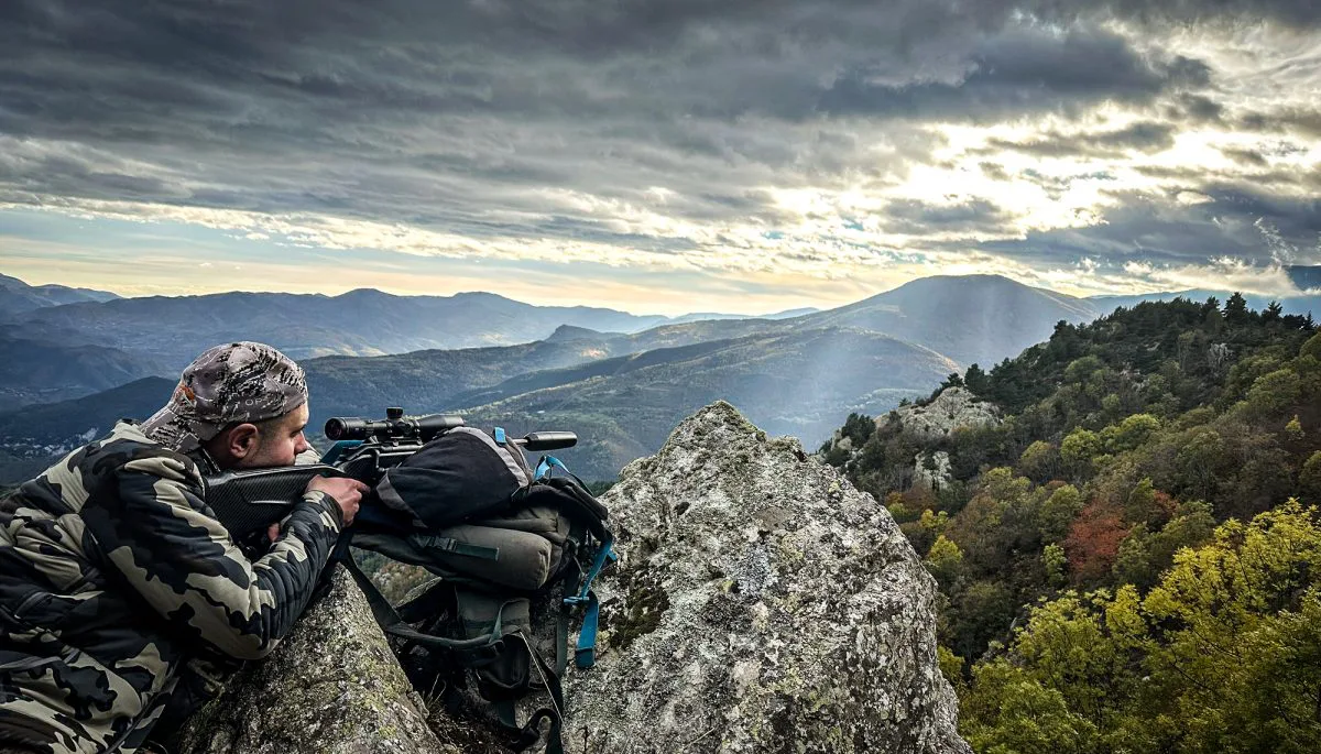Un individu camoufl&eacute; est accroupi sur un point de vue rocheux, scrutant &agrave; travers une paire de jumelles un vaste paysage montagneux sous un ciel nuageux spectaculaire.