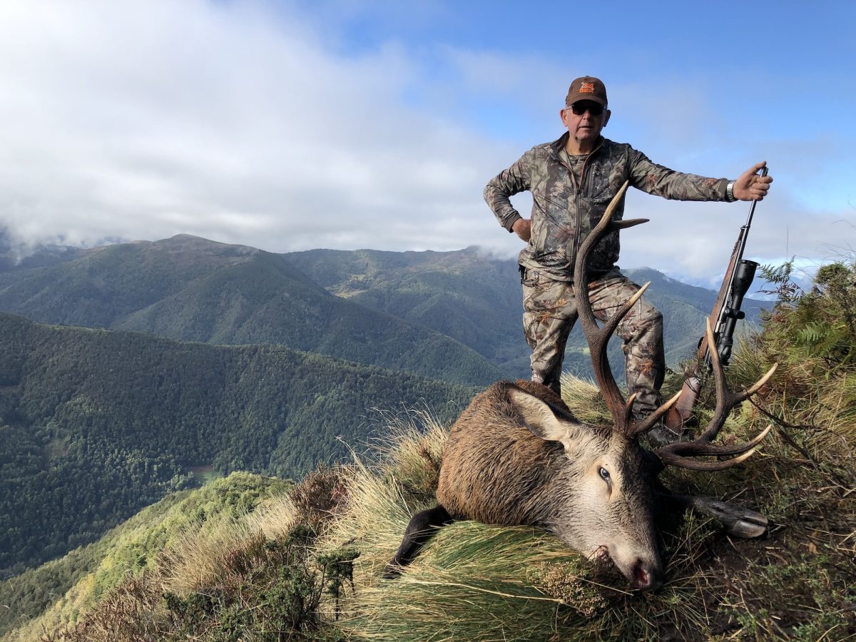 Un chasseur en tenue de camouflage posant avec son fusil à côté de l'accueil, un cerf abattu sur fond de collines boisées et un ciel partiellement nuageux.