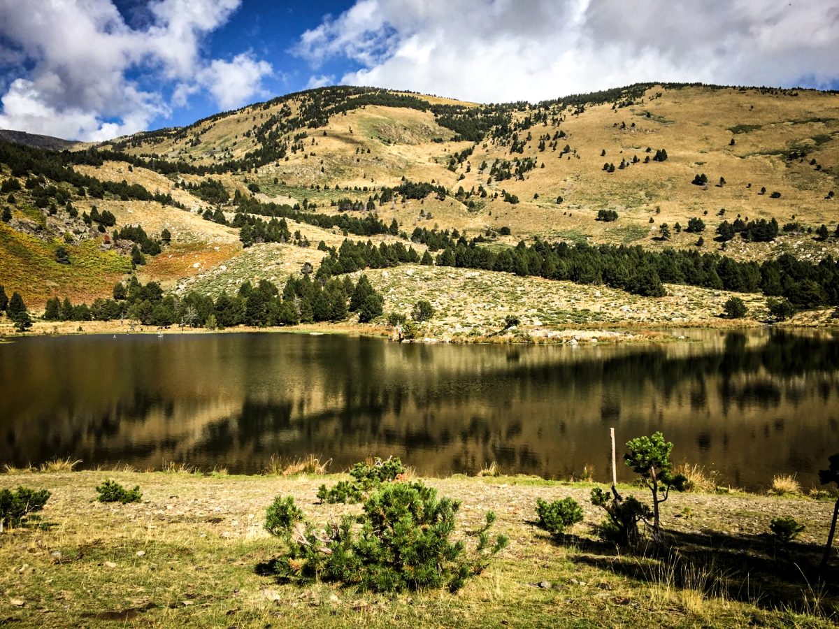 Tranquil mountain lake with reflections of towering hills and sparsely vegetated slopes under a partly cloudy sky.