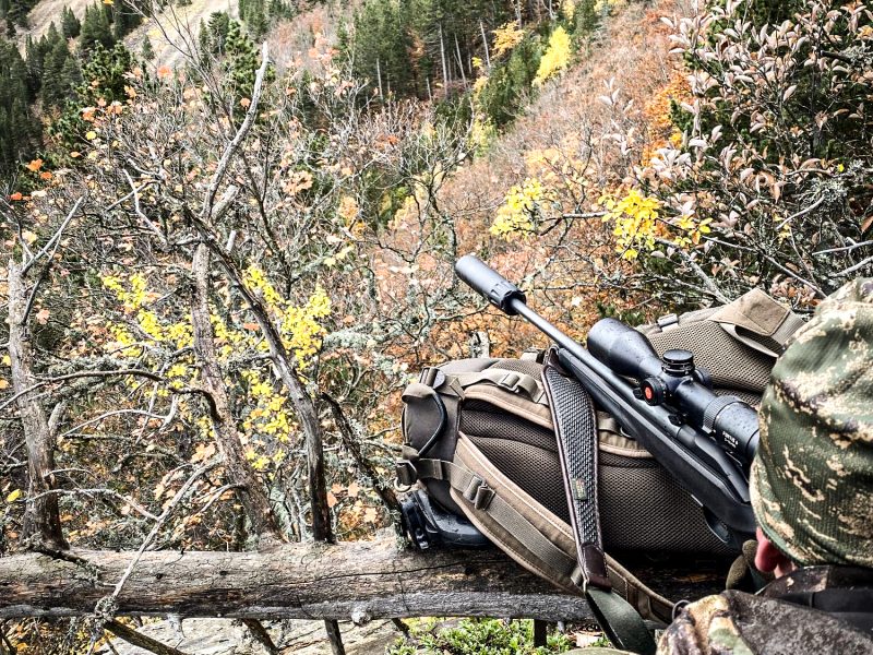 A hunter in camouflage gear rests with a rifle atop a sturdy rucksack, overlooking a dense autumn forest from a high vantage point.