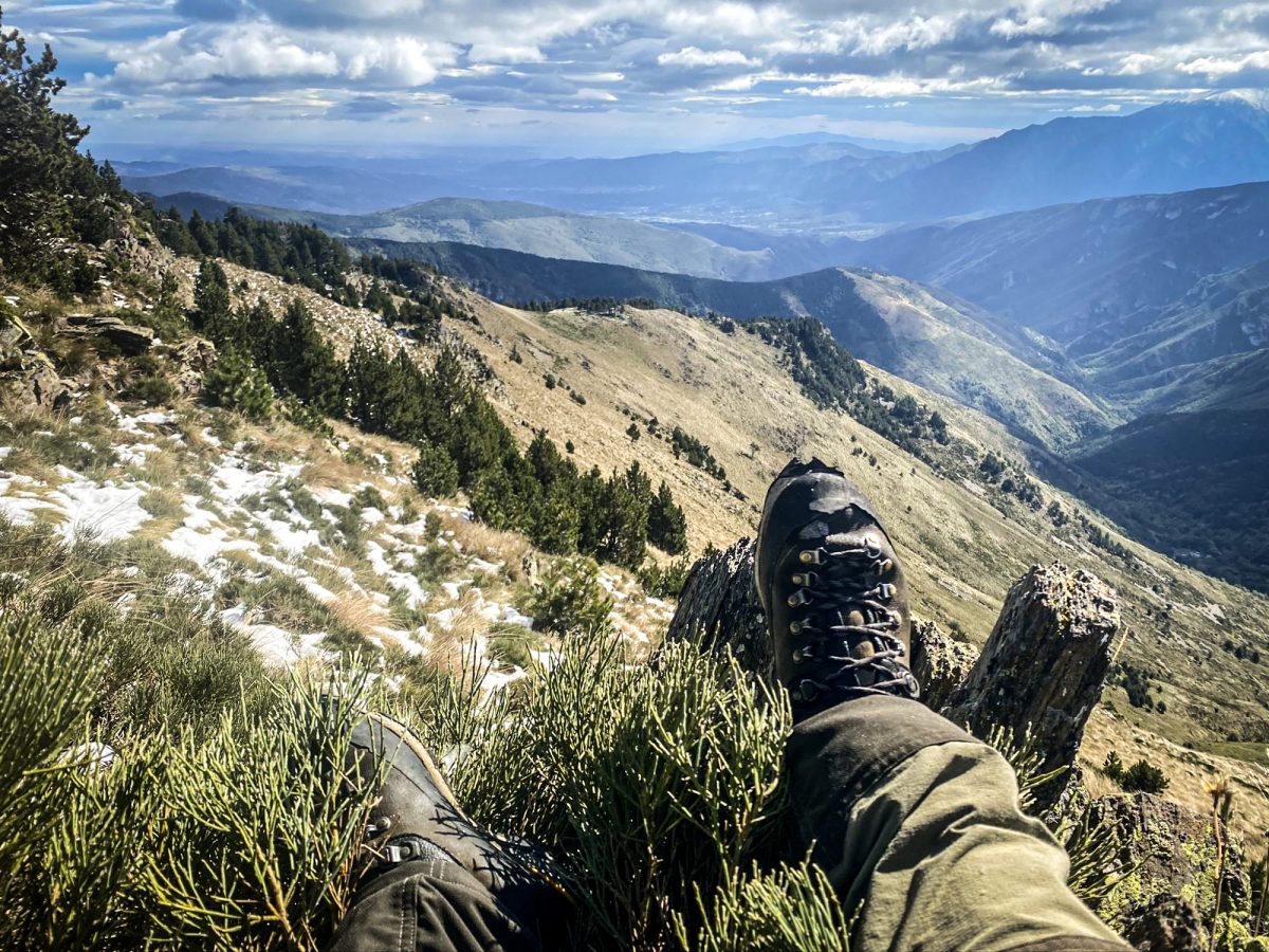 Los pies del cazador descansan en el borde de una montaña, con una vista impresionante del valle y los picos lejanos bajo un cielo inmenso.