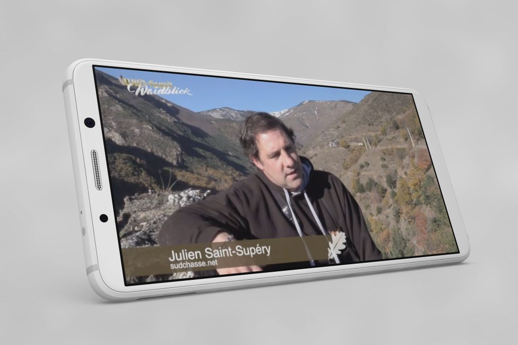 Un teléfono inteligente colocado de lado que presenta una entrevista en vídeo con Julien Saint-Supéry, frente a majestuosas montañas, se identifica por un texto superpuesto que indica 