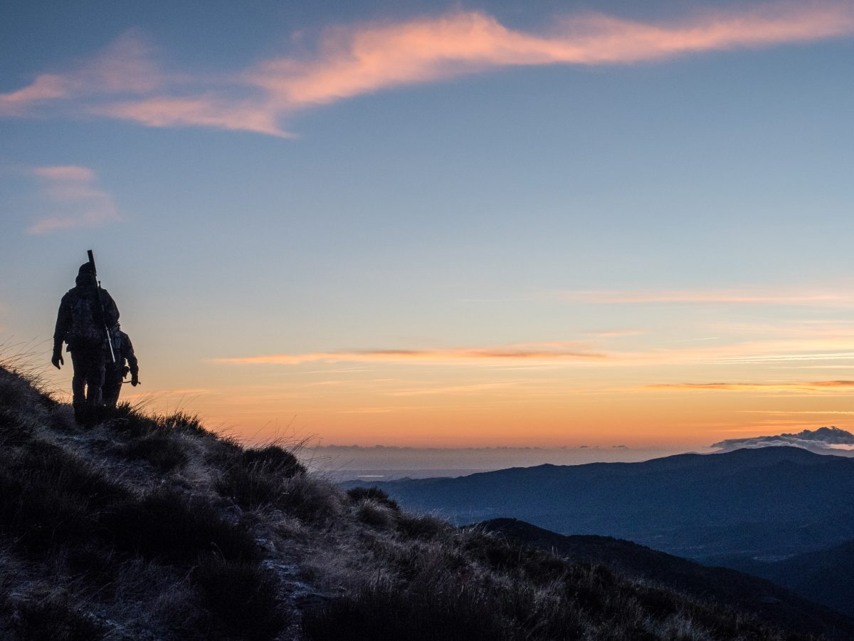 A hunter climbing a mountain trail at dusk, with a gradient of sunset colors painting the sky in the background.