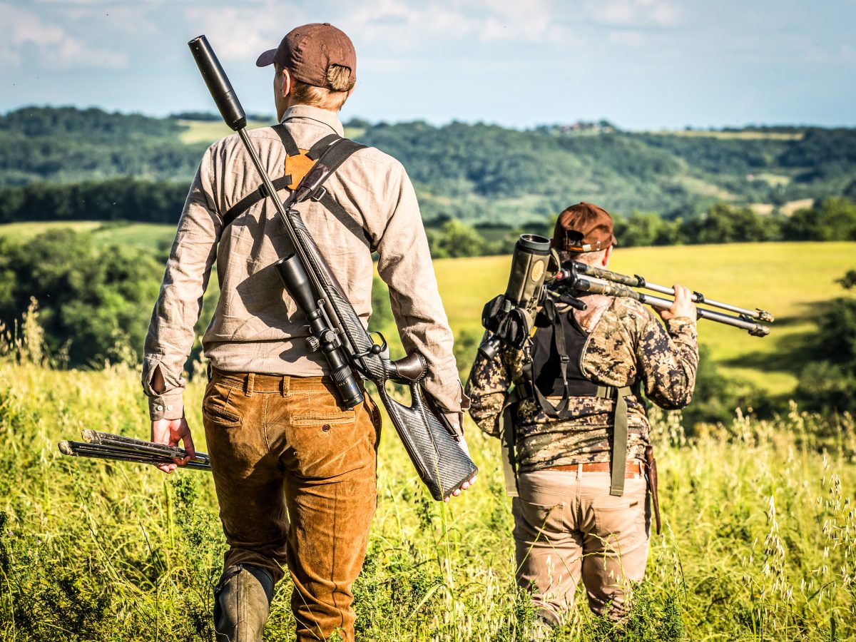 Deux individus en tenue de plein air, un fusil sur l'épaule, marchant dans un champ herbeux, apparemment en excursion de chasse.