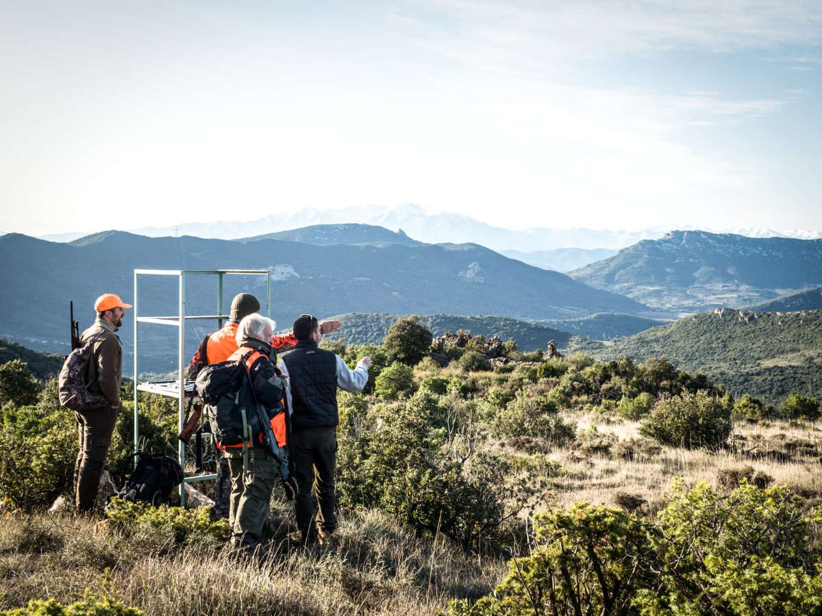 Un grupo de cazadores con mochilas se detienen para admirar la vista de un paisaje montañoso desde un mirador cubierto de hierba.