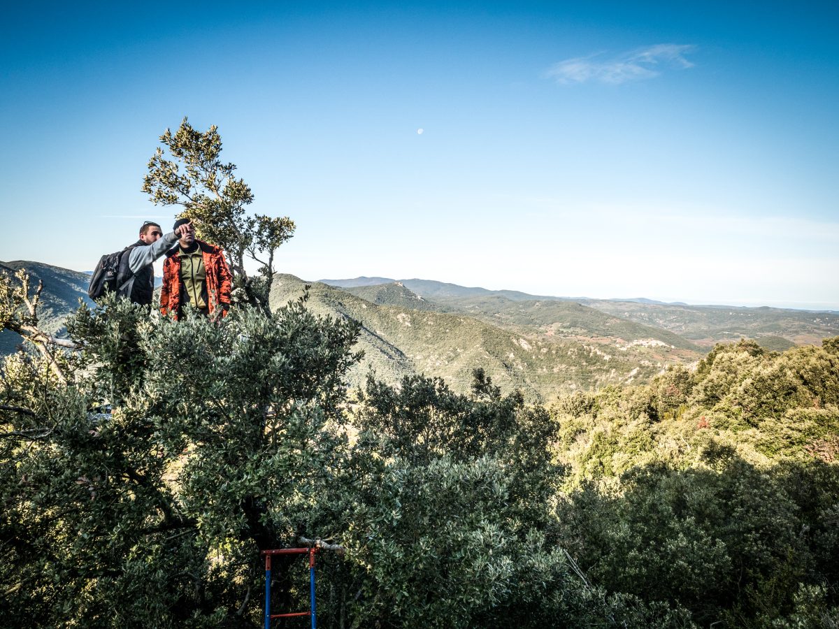 Dos cazadores en la cima de una montaña, deleitándose con la vasta vista panorámica con la luna visible en el cielo a plena luz del día.
